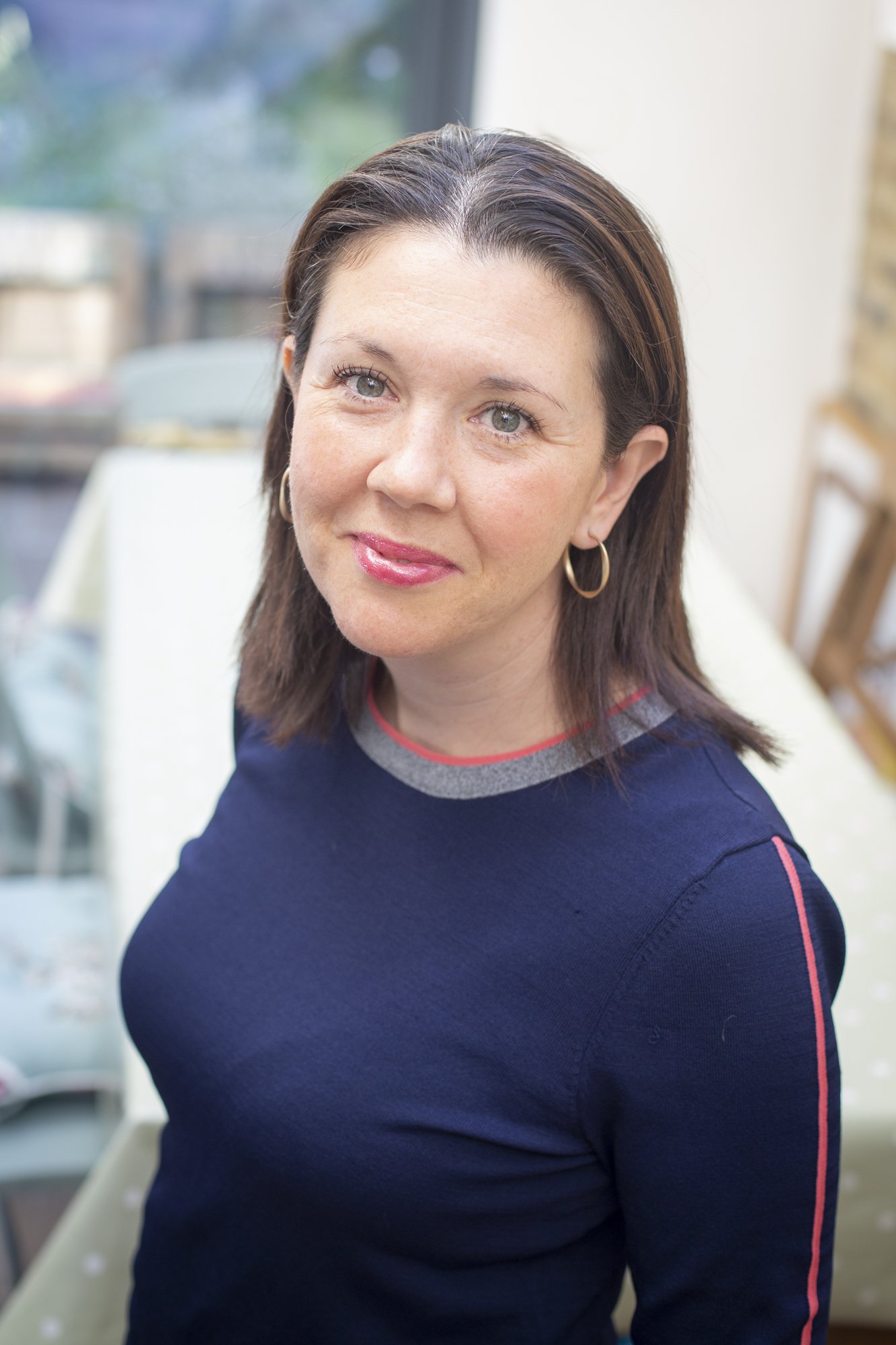 A woman with shoulder-length brown hair, gold hoop earrings, wearing a navy blue top with pink and gray accents, smiling and looking at the camera indoors.