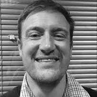 Black and white photo of a smiling man with short, wavy hair in front of a window with blinds.