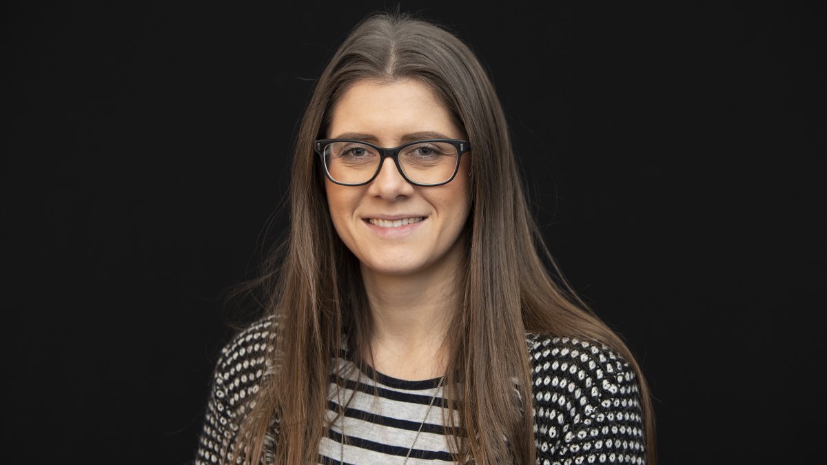 A woman with long brown hair, glasses, and a striped shirt, smiling against a black background.