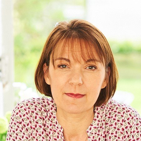 A woman with short brown hair and wearing a pink patterned shirt, sitting indoors with a blurred green outdoor background.
