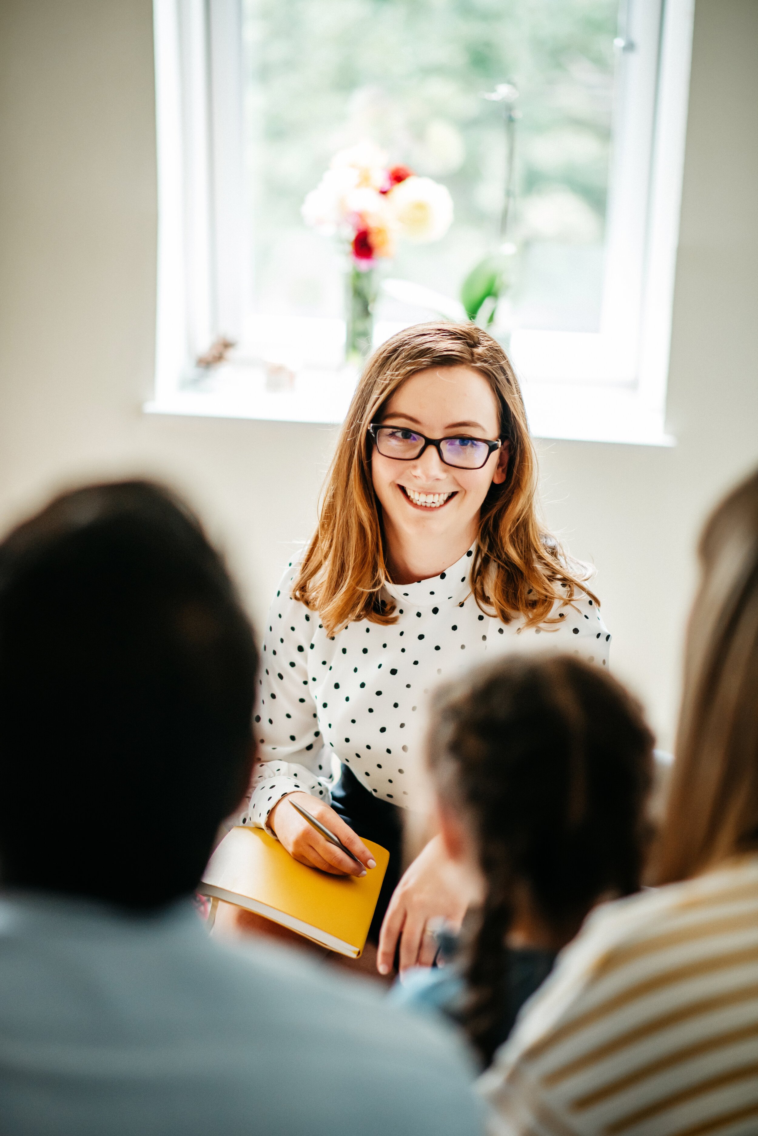 A woman with glasses, shoulder-length brown hair, and a white polka-dot shirt sitting with a notepad, talking to a group of people in a room with a window and a vase of flowers in the background.