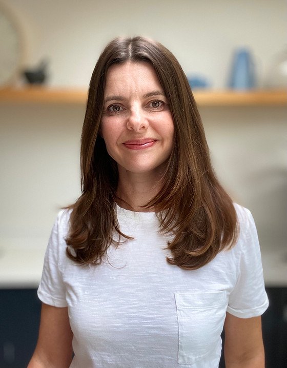 A woman with long brown hair wearing a white t-shirt, smiling in an indoor setting with blurred background shelves.