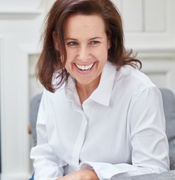 A woman with brown hair smiling and wearing a white shirt, sitting indoors.