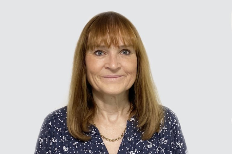 A woman with shoulder-length reddish-brown hair, wearing a dark blue blouse with white floral patterns and a gold necklace, smiling at the camera against a plain light gray background.