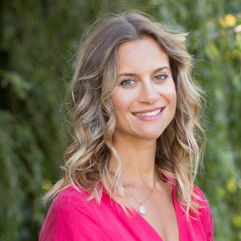 A woman with wavy blonde hair smiling outdoors, wearing a pink top and a silver necklace with a round pendant.