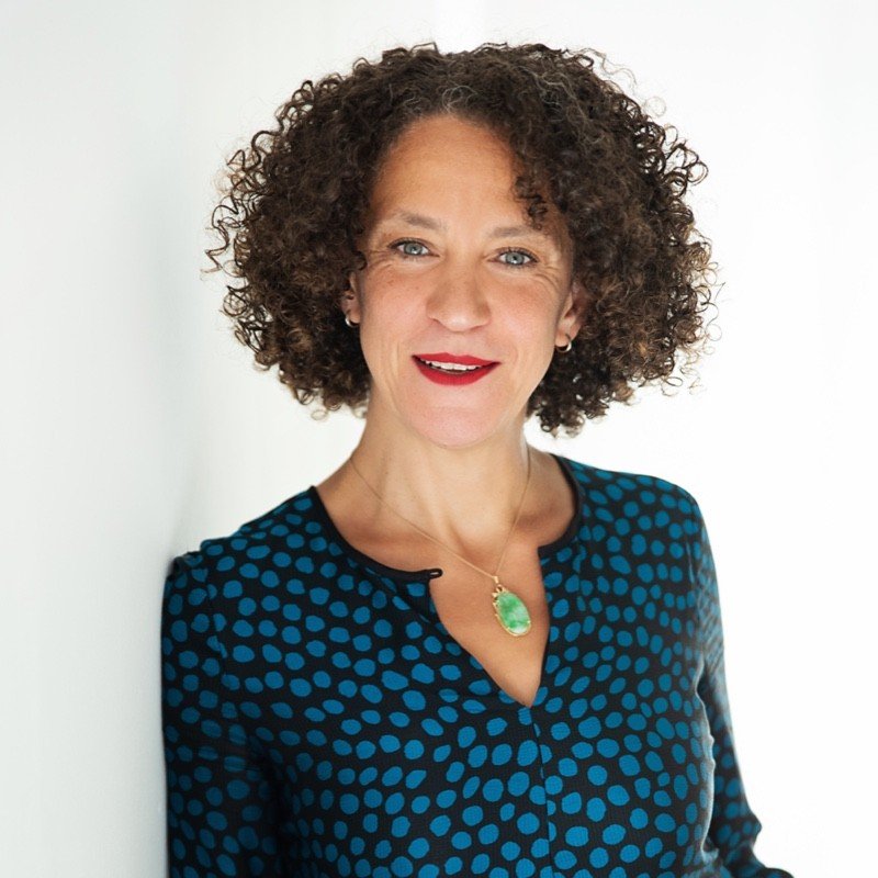 A woman with curly brown hair wearing a navy blue dress with blue polka dots and a green pendant necklace, smiling against a white background.