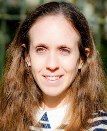 A woman outdoors with long wavy brown hair, wearing a navy and white striped shirt, smiling at the camera.