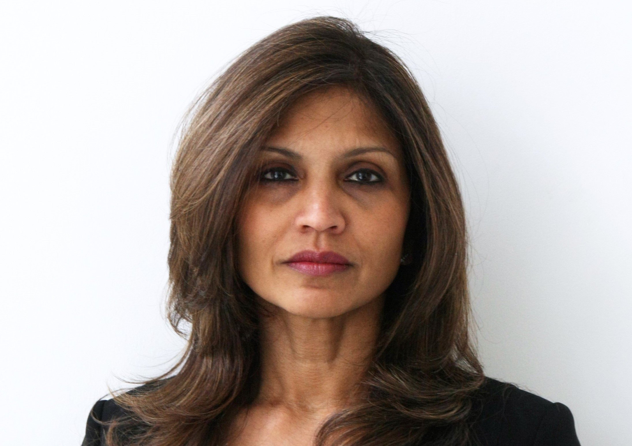 A middle-aged woman with long brown hair and light skin, wearing a black top, standing against a plain white wall.