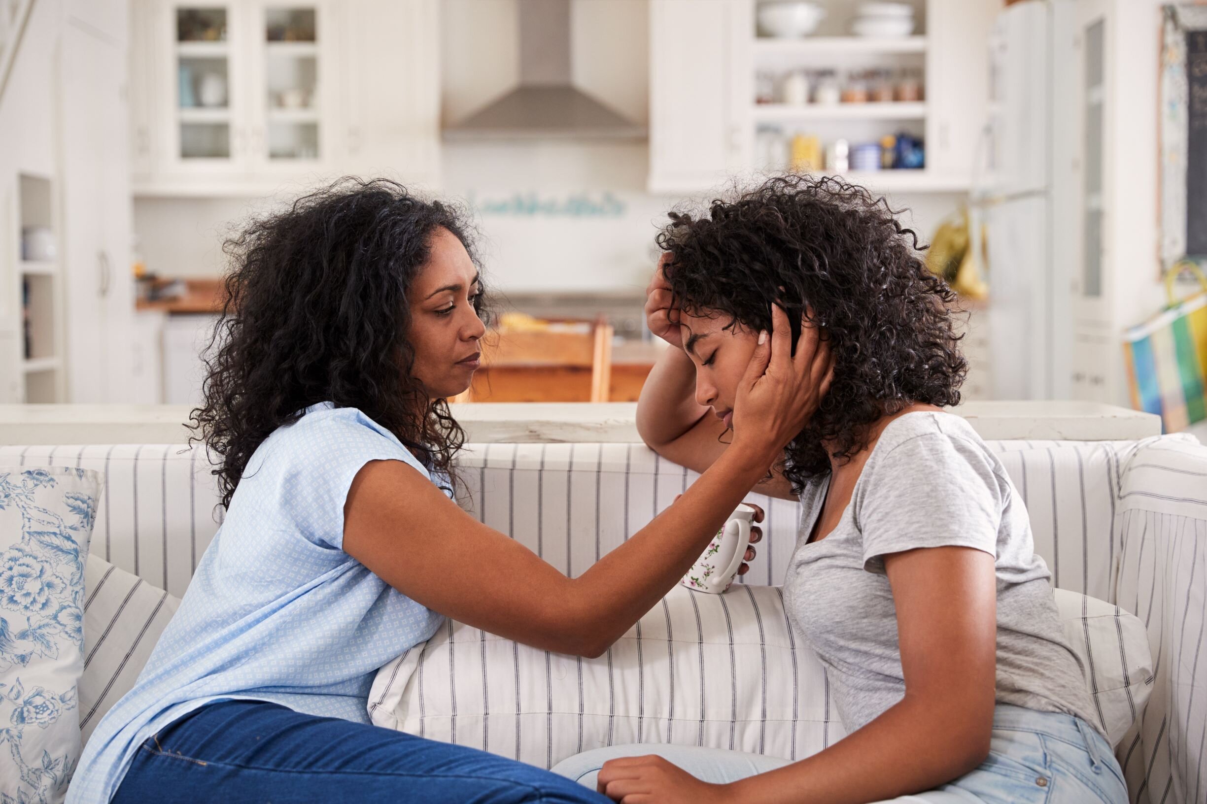 A woman comforts a distressed young girl in a domestic setting, holding her head and shoulder.