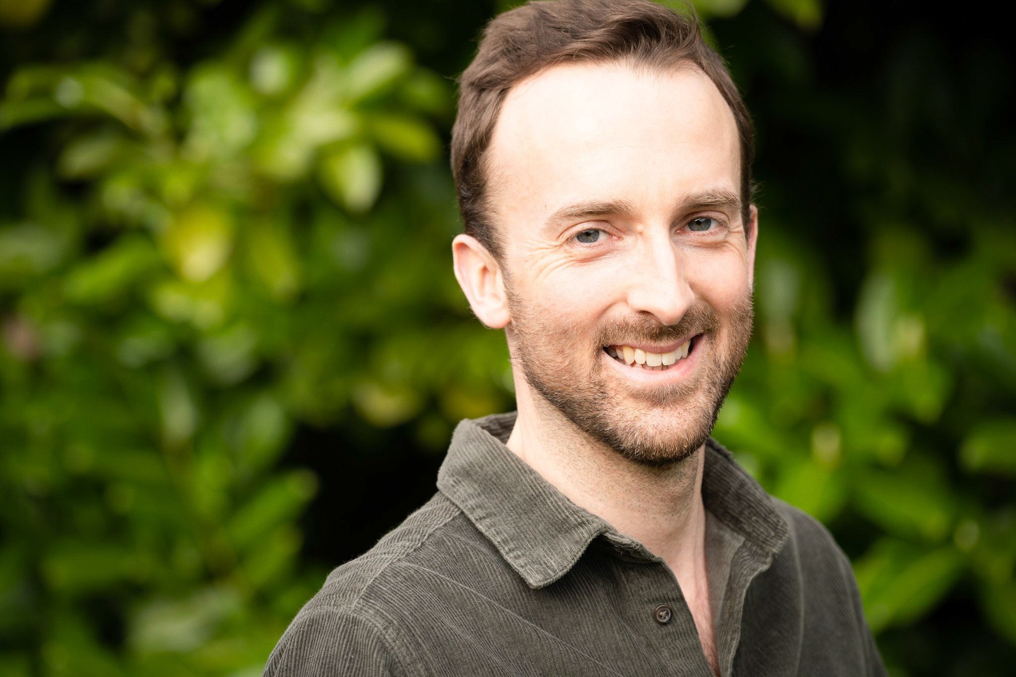 Smile of a man with a beard and brown hair outdoors with green foliage in the background.