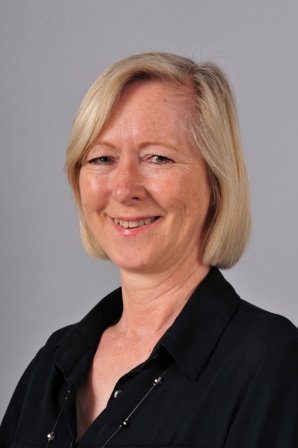 A smiling middle-aged woman with blonde hair, wearing a black collared shirt, against a gray background.
