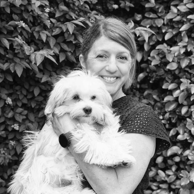 A woman smiling and holding a fluffy small dog in front of leafy bushes.