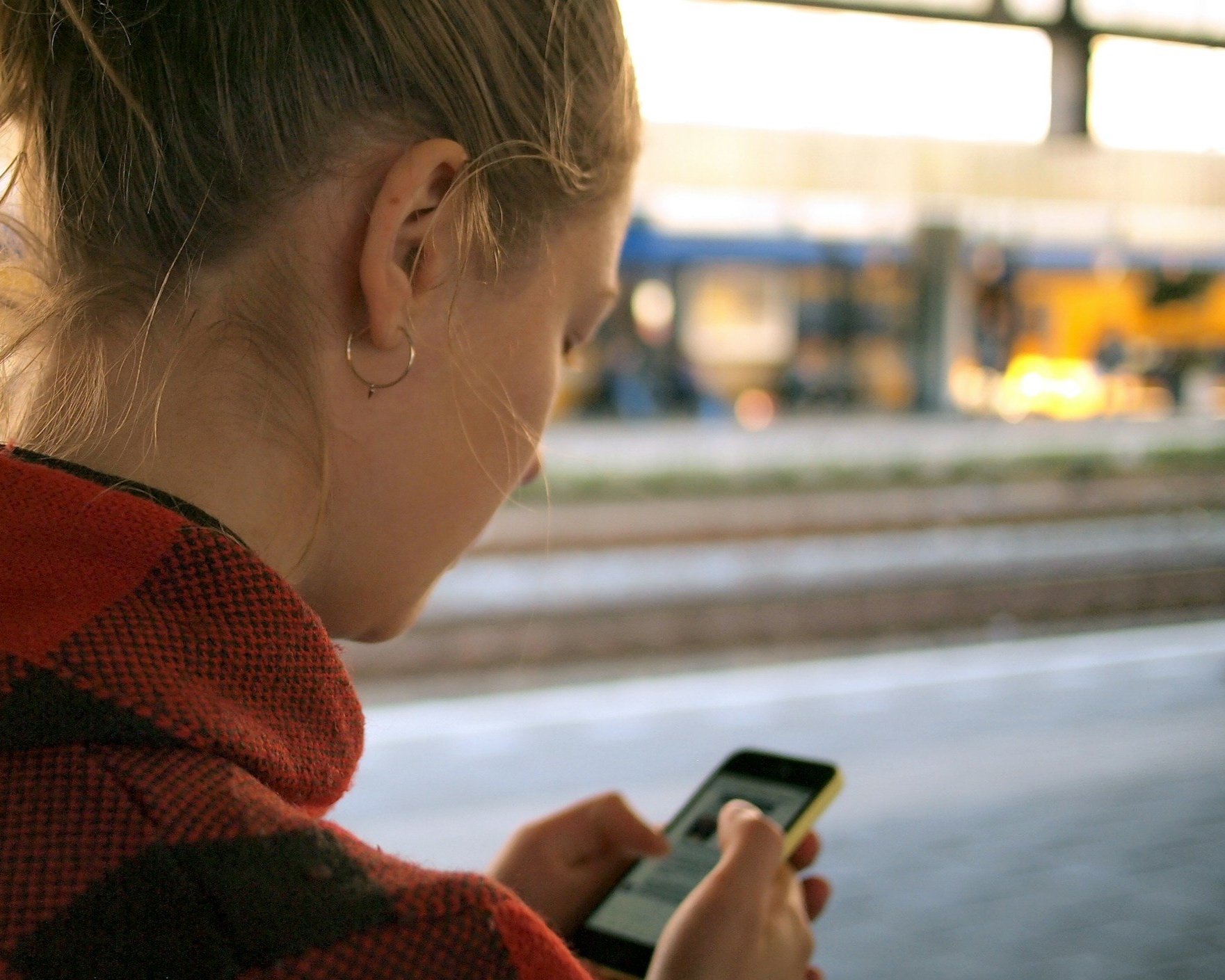A woman with blonde hair tied back, wearing hoop earrings and a red and black checkered jacket, is looking at her phone with a blurred outdoor industrial background.