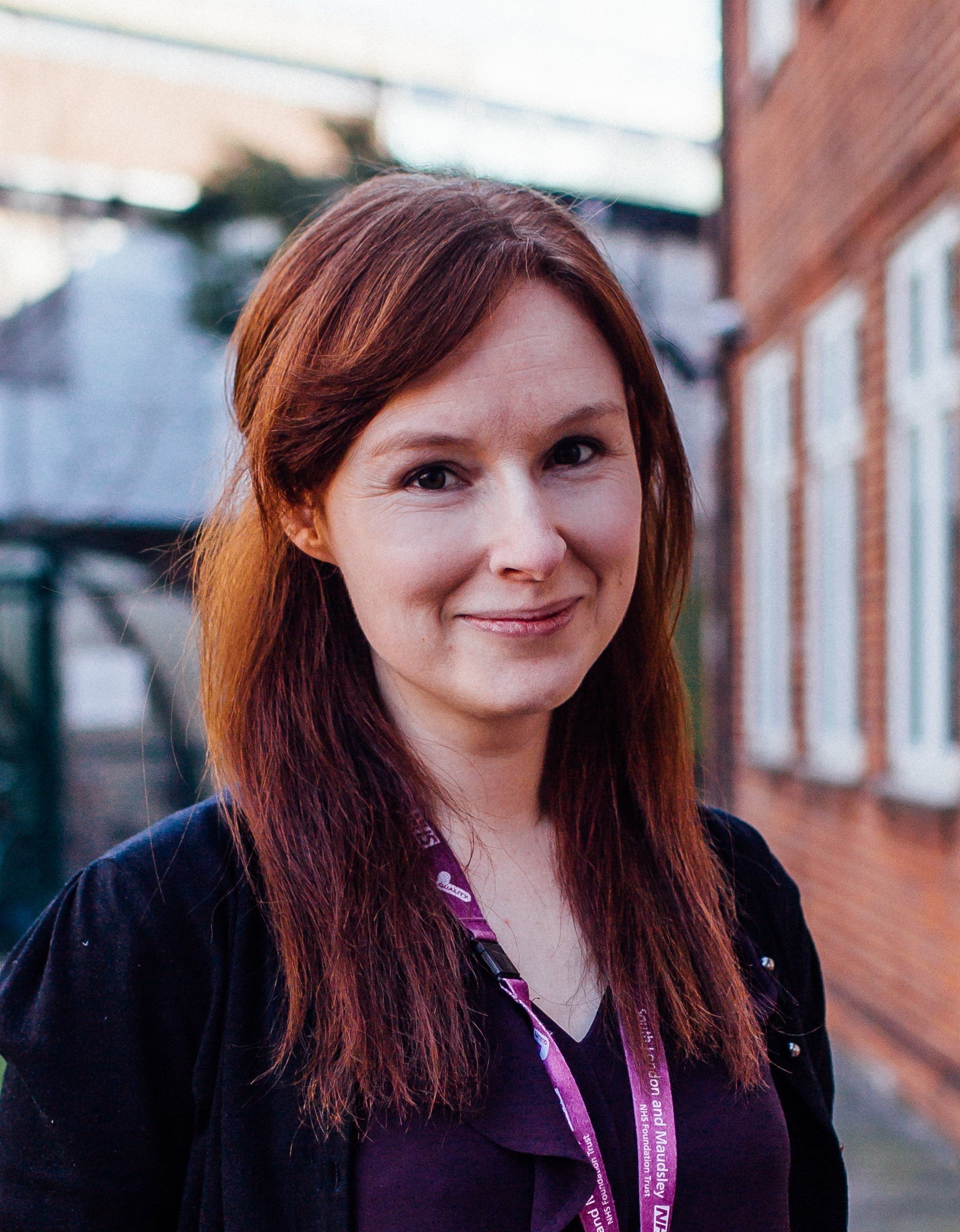 A woman with shoulder-length reddish-brown hair and a slight smile, standing outdoors in a residential neighborhood with brick buildings and a cloudy sky in the background.
