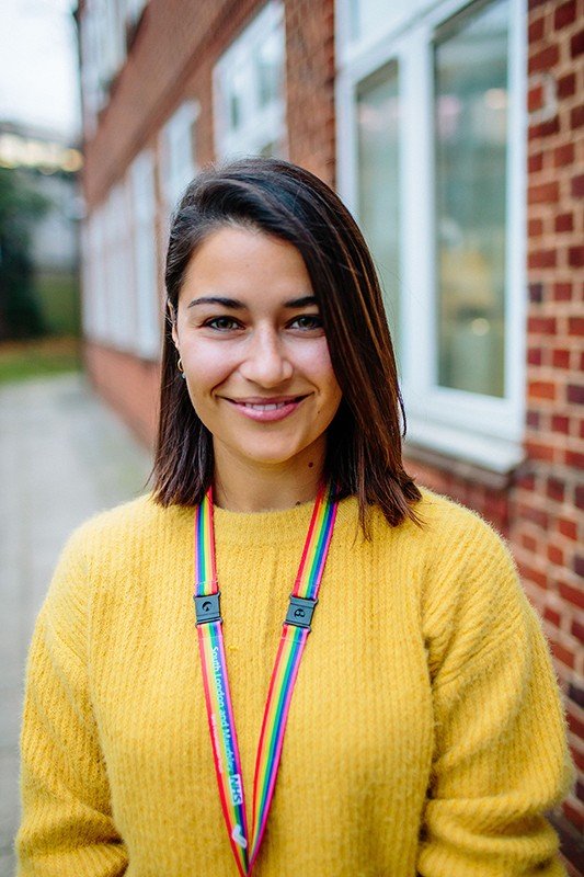 Young woman with dark hair wearing a yellow sweater and a rainbow-colored lanyard, standing outdoors near a brick building with windows.
