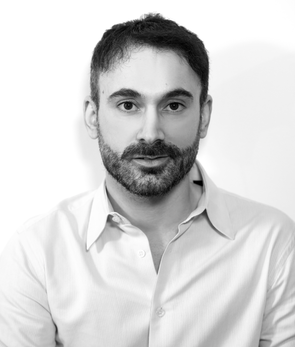 Black and white portrait of a man with a beard and short dark hair, wearing a light-colored collared shirt, looking directly at the camera.