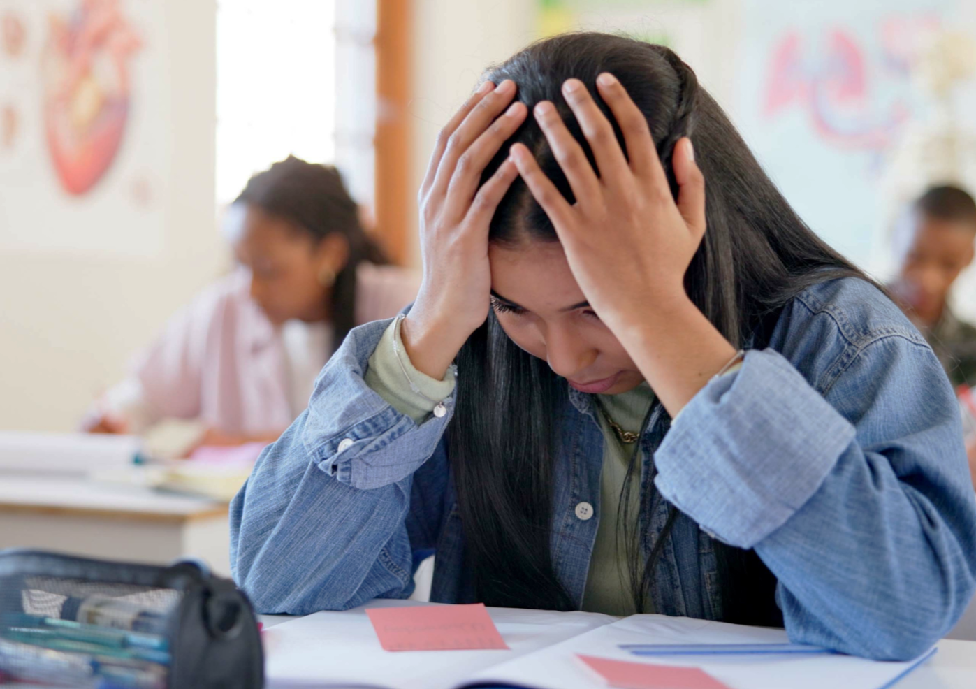A young woman sitting at a desk with her hands on her head, appearing stressed or overwhelmed with schoolwork in a classroom setting.