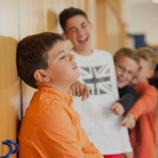 A young boy leaning against a school wall, eyes closed, with three smiling classmates in the background.